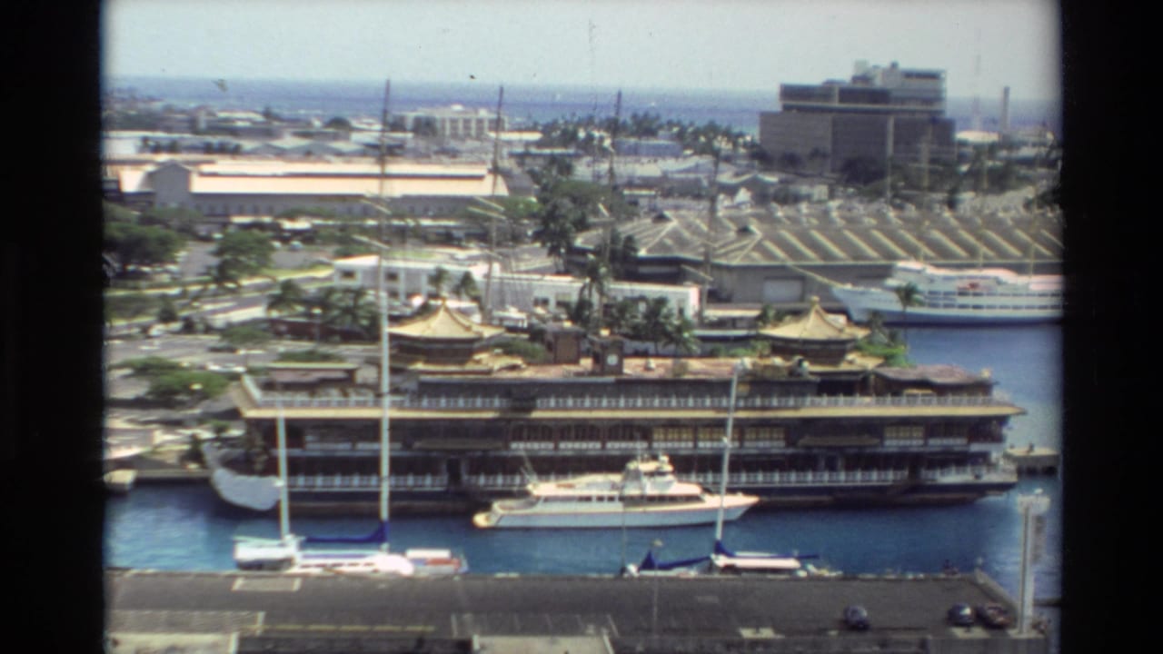 Wide harbor view with a large ship at a pier, sailboats, golden-roofed buildings, and city structures beyond.