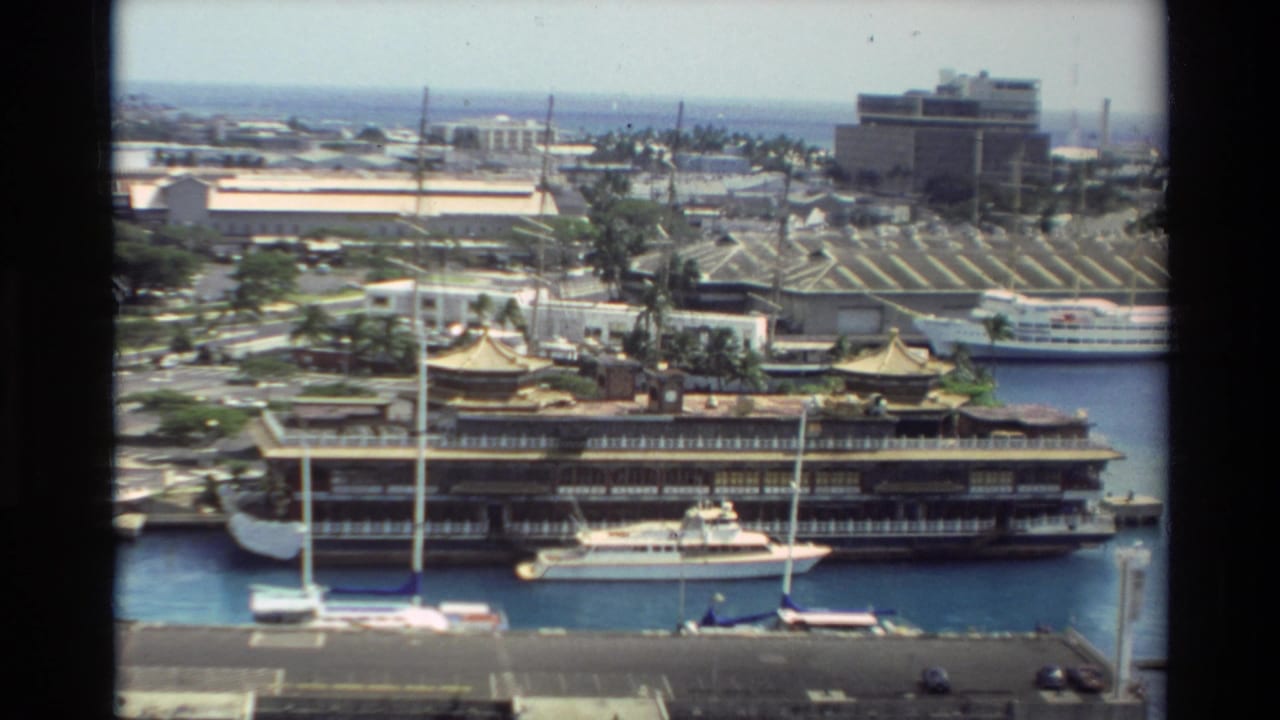 High-angle 1981 Hawaii harbor shot with a large vessel at the pier, nearby sailboats, palm trees, and waterfront buildings under bright daylight.