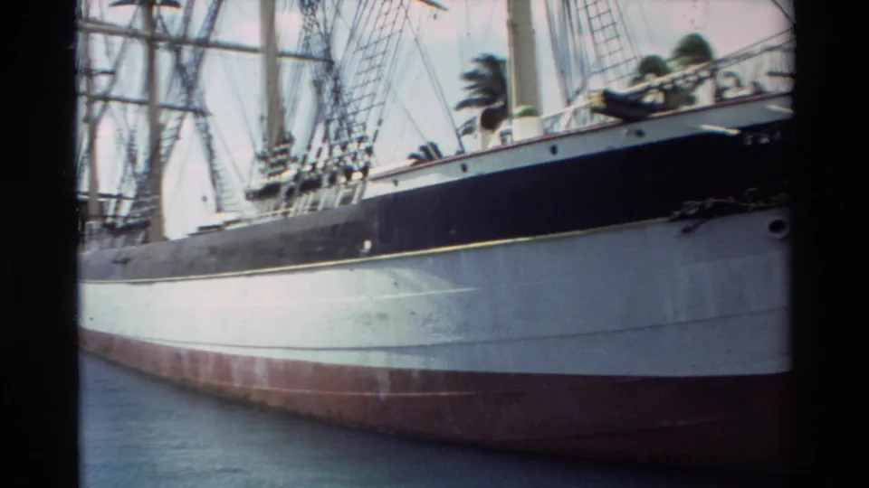 1981 Hawaii 8mm frame showing the side of a docked sailing ship, with white hull, dark band, red bottom, rigging, and calm water.