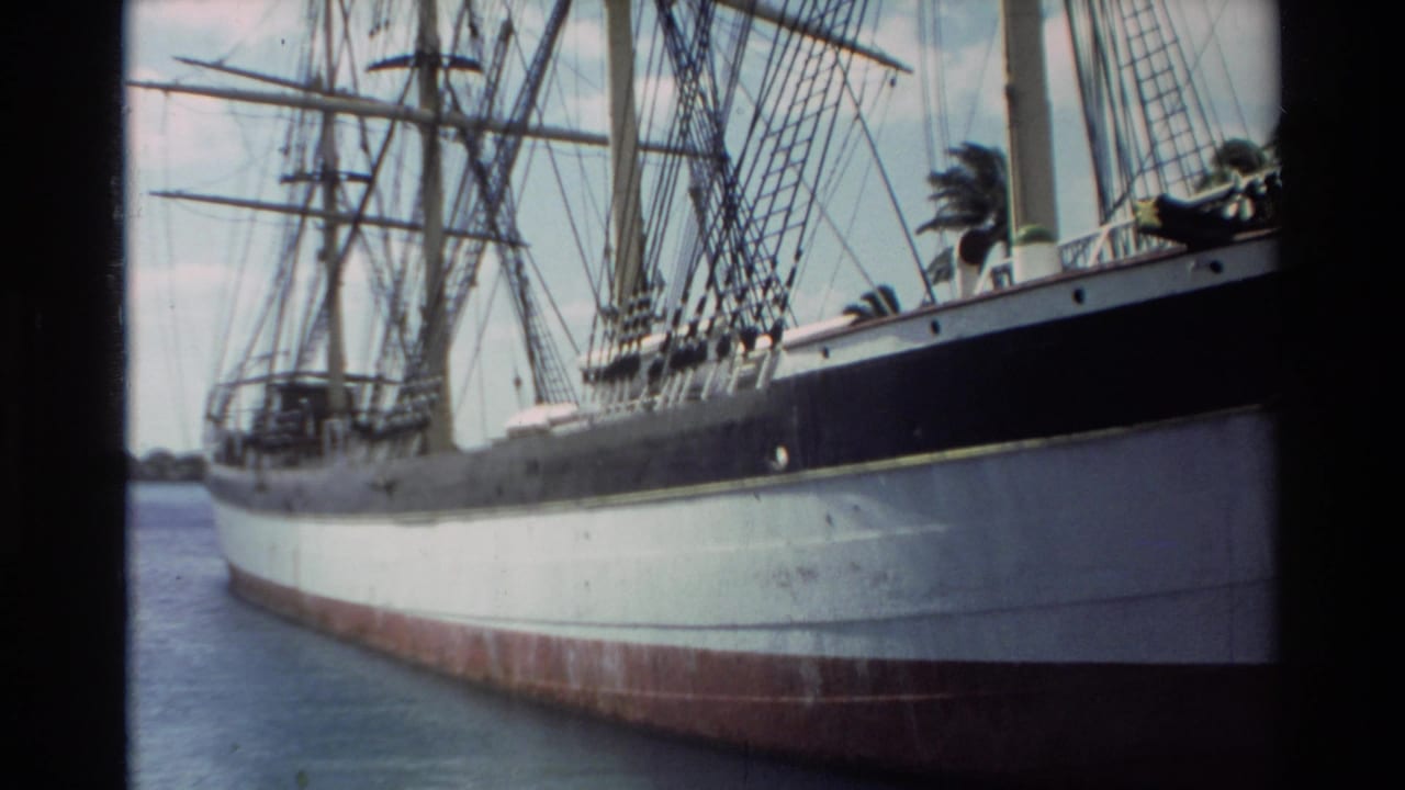 1981 Hawaii home-movie frame of a docked sailing ship seen from the side, with rigging above the hull and calm harbor water below.