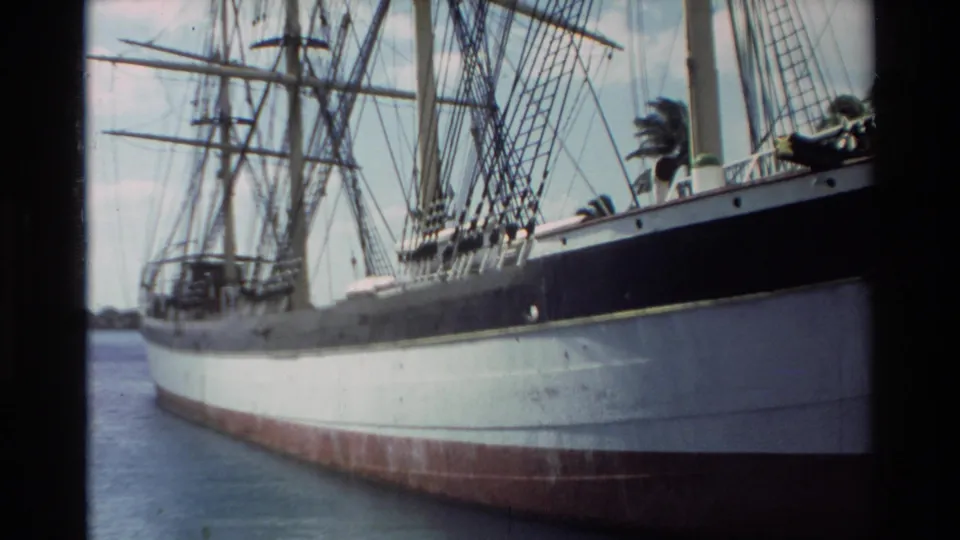 1981 Hawaii home-movie frame of a docked sailing ship seen from the side, with rigging above the hull and calm harbor water below.