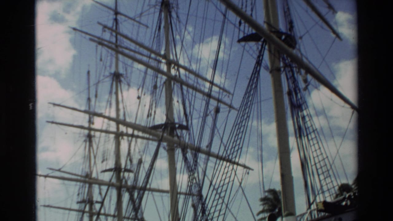 1981 Hawaii home-movie frame showing tall ship masts, rigging, and spars viewed from below against scattered clouds.