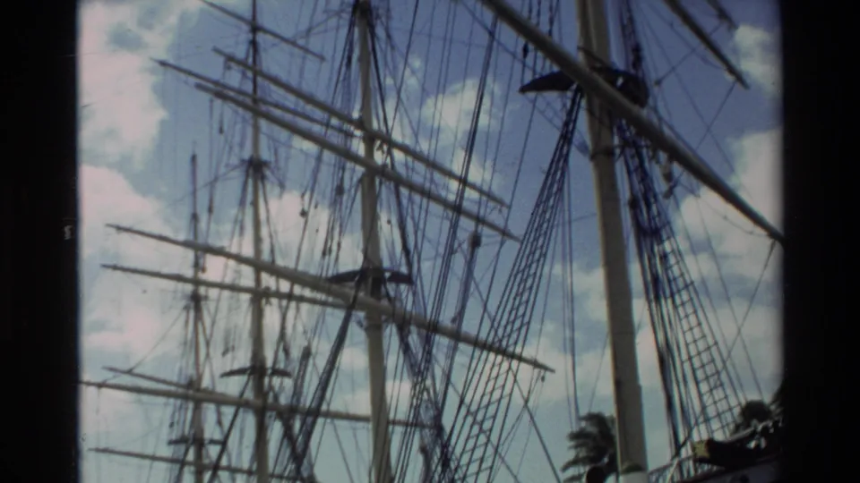 1981 Hawaii home-movie frame showing tall ship masts, rigging, and spars viewed from below against scattered clouds.