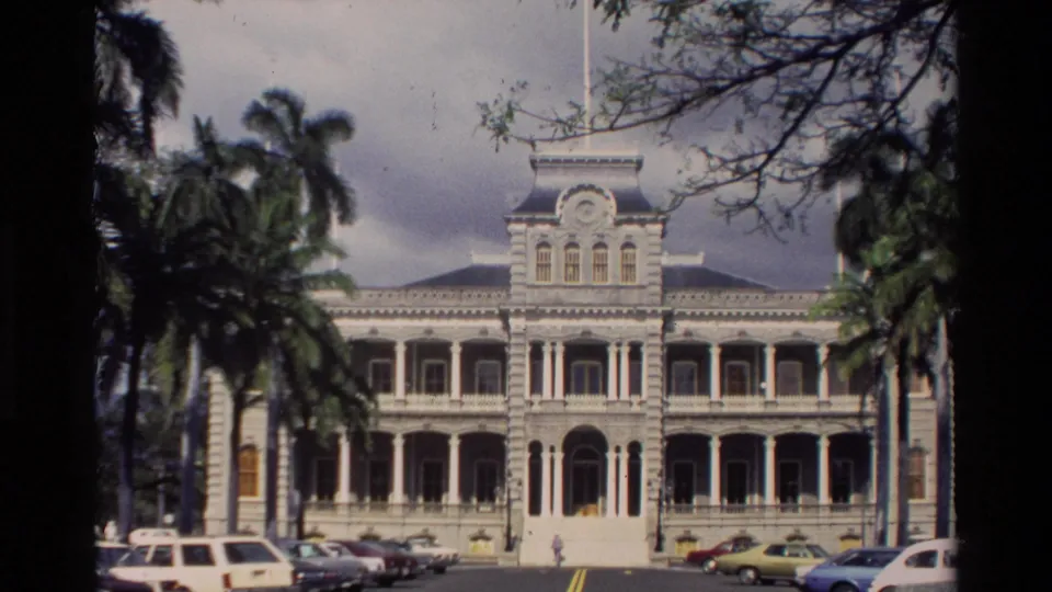 1981 Hawaii frame of a large white columned building framed by palm trees, with early-1980s cars parked in front and small people near the entrance.