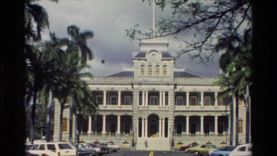 Static Hawaii travel view of the white historic-style building facade, palm trees, parked cars, and small figures near the front walkway.