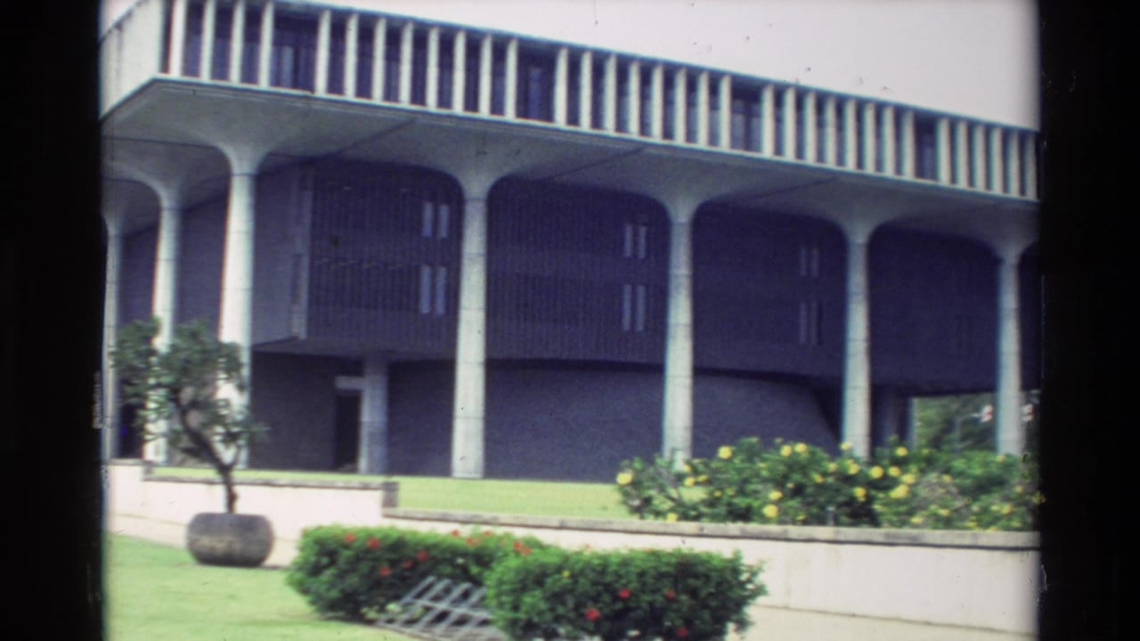 1981 Hawaii 8mm frame showing a modernist exterior with concrete columns, landscaped beds, lawn, shrubs, and a small tree.