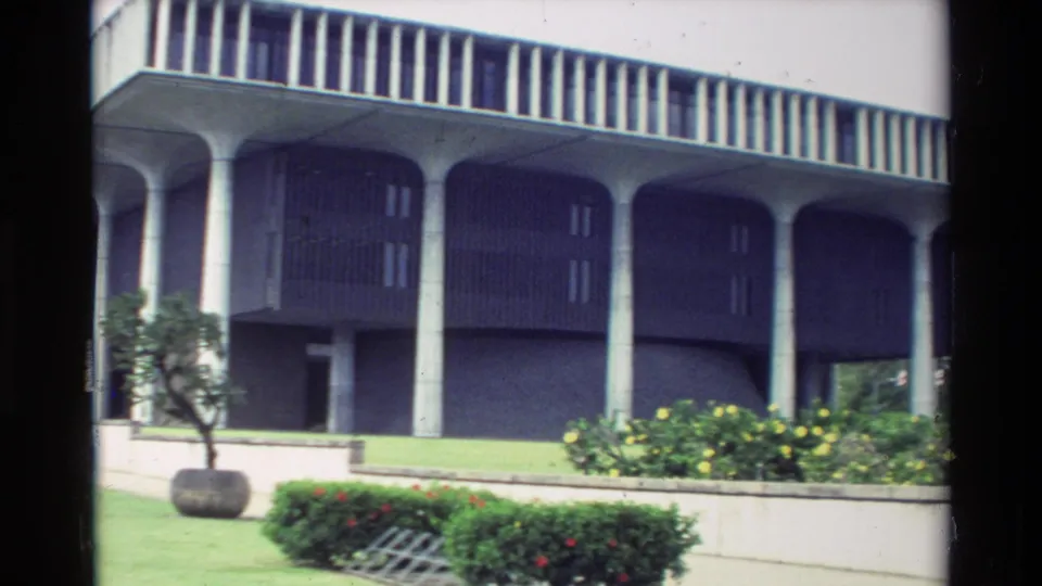 1981 Hawaii 8mm frame showing a modernist exterior with concrete columns, landscaped beds, lawn, shrubs, and a small tree.