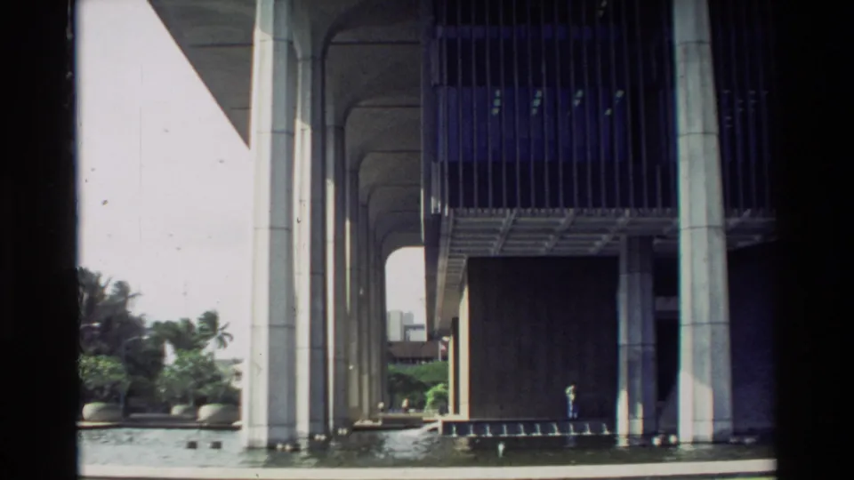 1981 Hawaii frame looking beneath the raised modernist structure, with columns, vertical screens, water, palms, and a small distant person.