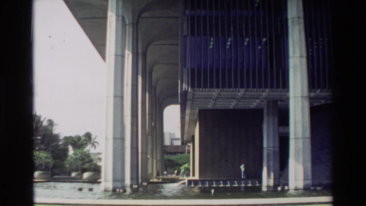 Modernist concrete building over water with palms and a small person near the shaded walkway.