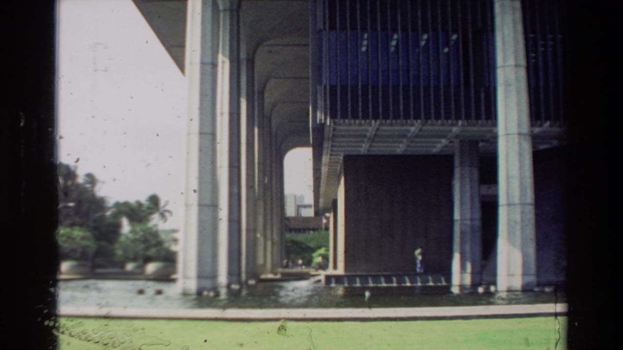 Wide 8mm view of a Hawaii modernist building with massive columns, water in the foreground and a distant person under shade.