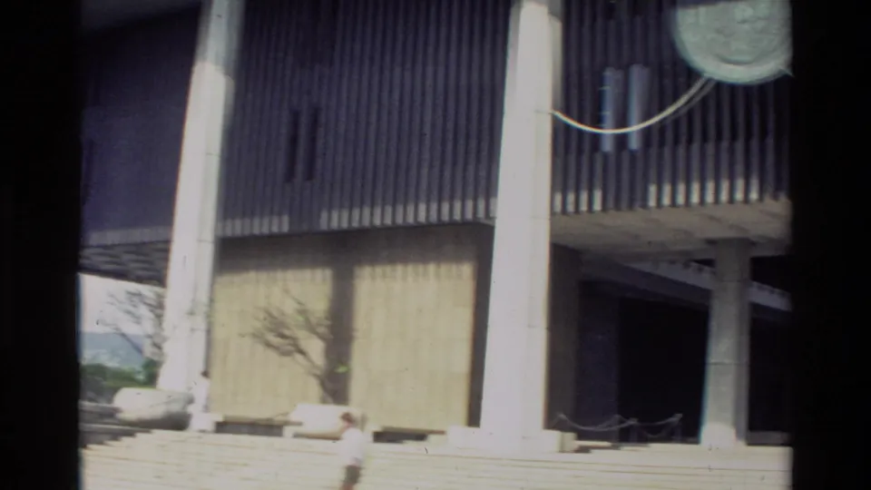 Closer 1981 Hawaii home-movie frame showing vertical facade screens, concrete supports, broad steps and a cropped visitor in the foreground.