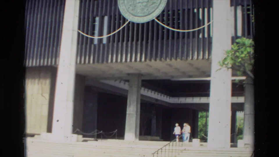 1981 Hawaii 8mm frame with two visitors near steps under a modernist concrete structure with screens, rope barriers and nearby greenery.
