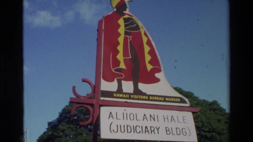 Home-movie close view of the Aliiolani Hale Judiciary Building marker with Hawaii Visitors Bureau artwork and trees behind it.