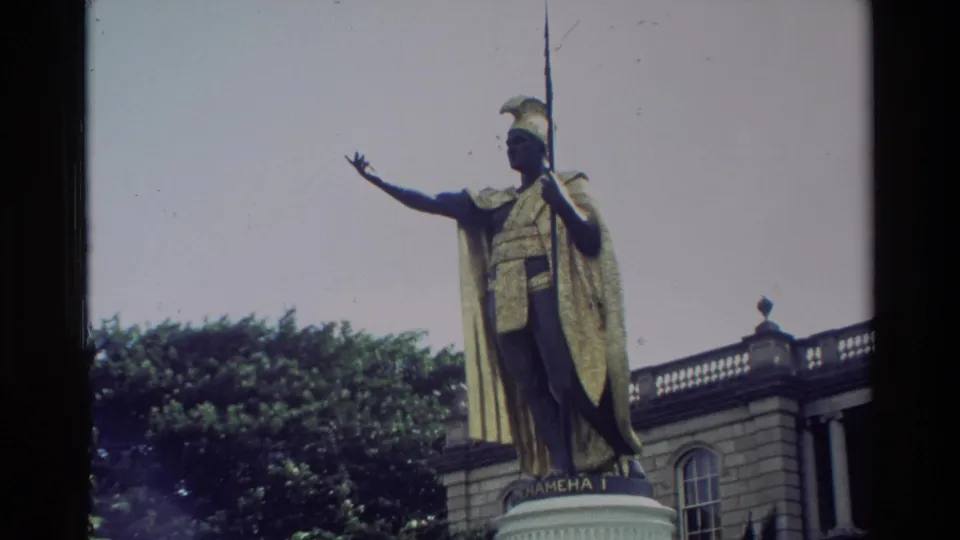 1981 Hawaii home-movie frame showing the King Kamehameha I statue, golden cloak, spear, pedestal, trees, and arched building.