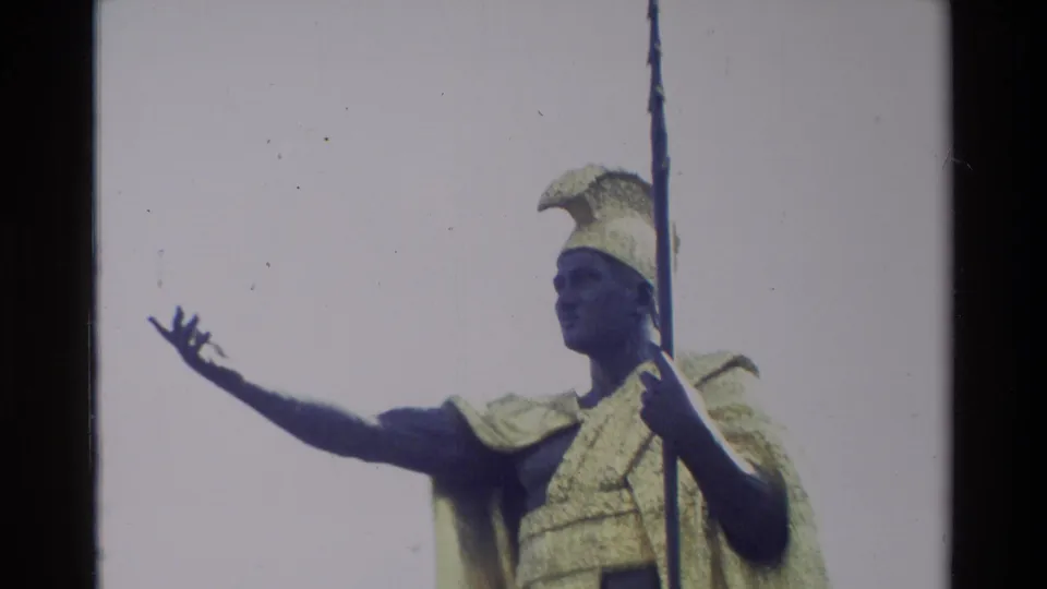 Closer 1981 Hawaii frame of the King Kamehameha I statue with feathered helmet, golden cloak, spear, and extended hand against pale sky.