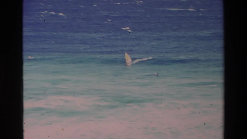 A windsurfer crosses turquoise Hawaii water in a grainy 1981 8mm home-movie frame.