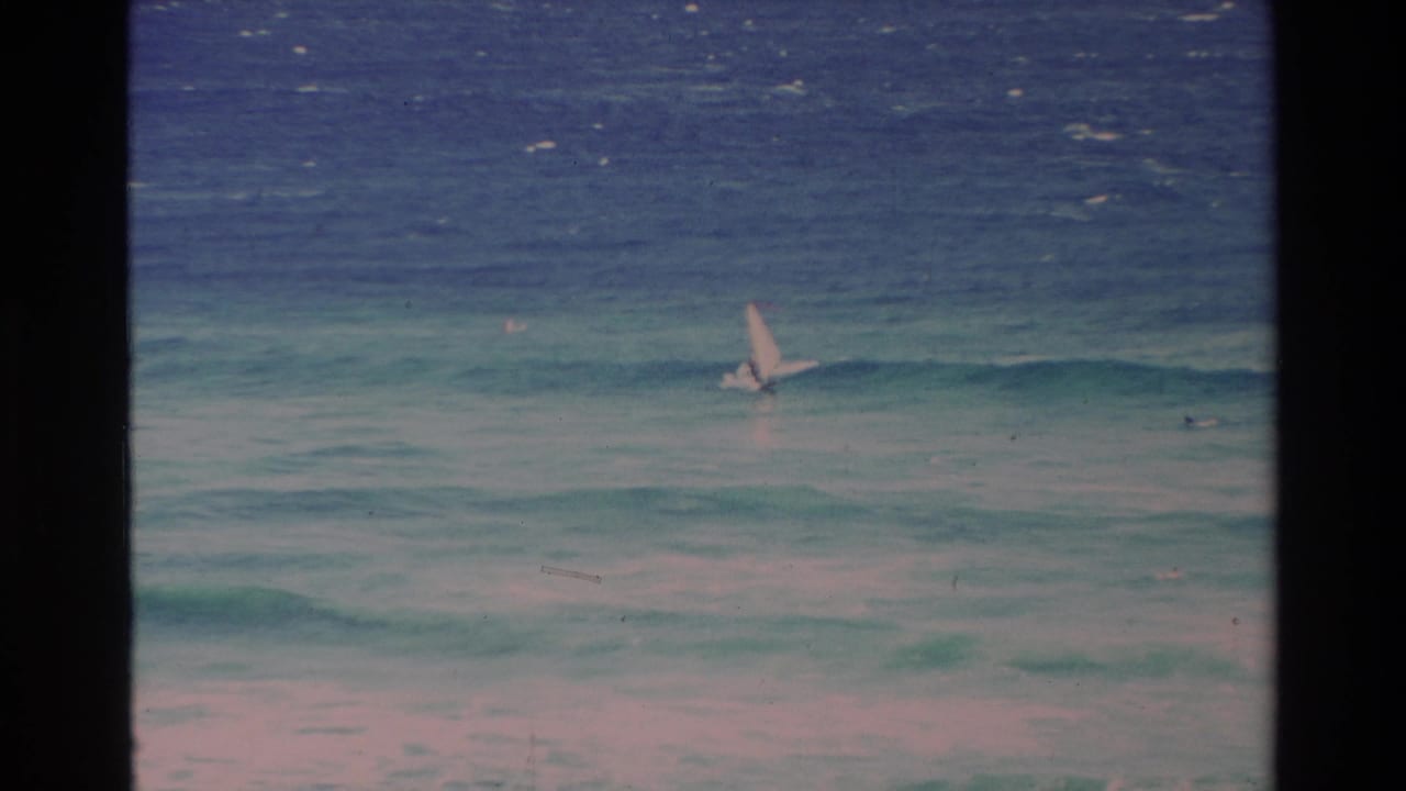 1981 Hawaii coastal frame with a small pale sail set against rolling turquoise surf and darker blue water beyond.