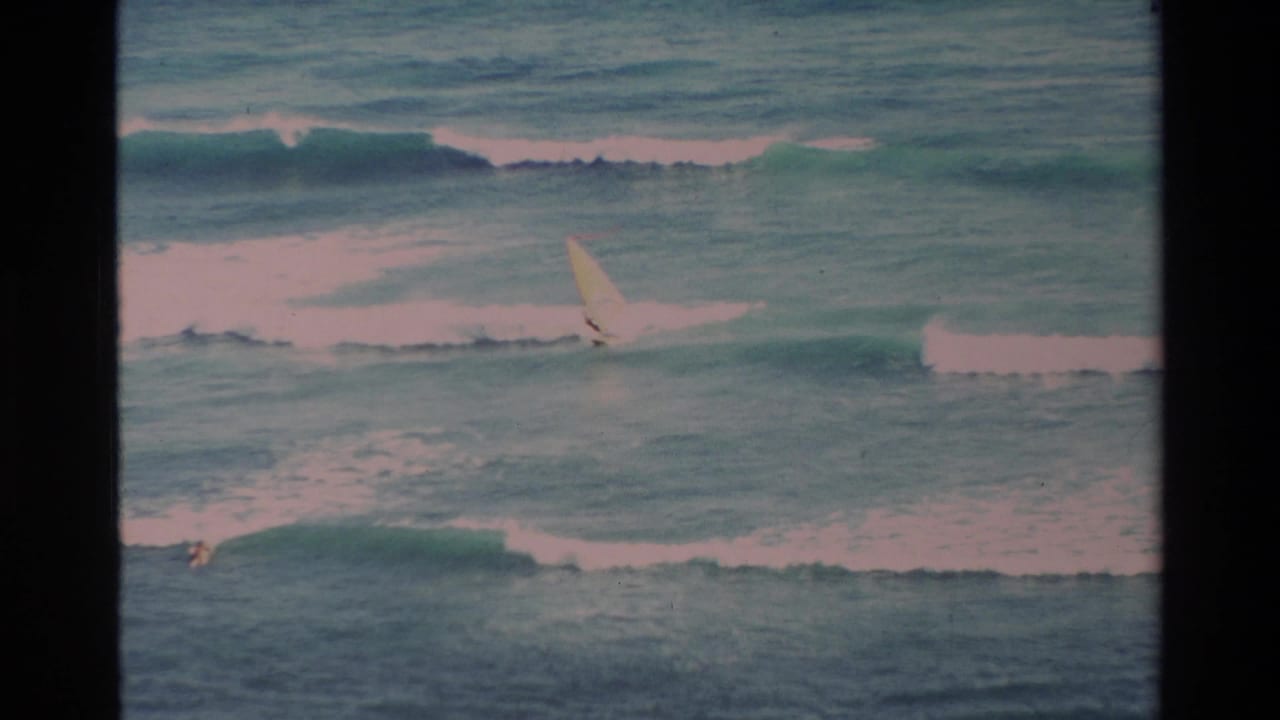 1981 Hawaii 8mm frame showing a distant windsurfer crossing turquoise surf with white waves breaking behind them.