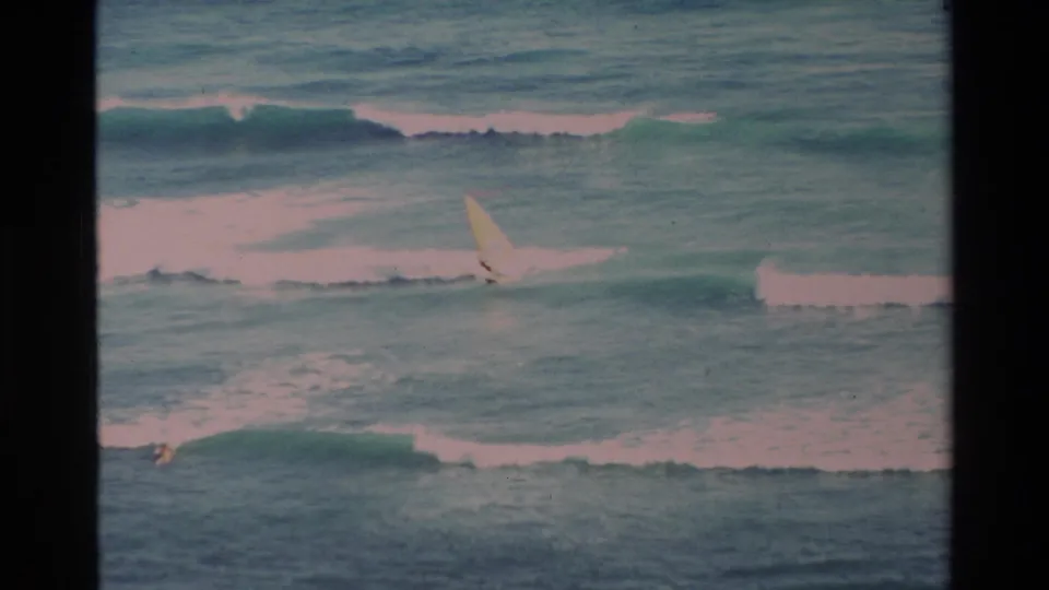 1981 Hawaii 8mm frame showing a distant windsurfer crossing turquoise surf with white waves breaking behind them.
