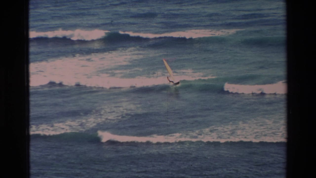 Color-faded 8mm view of a windsurfer off Hawaii, leaning into the sail while moving across rough blue-green water.