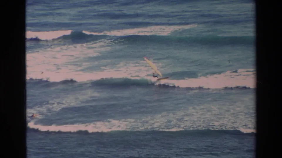 Distant 1981 Hawaii home-movie frame with a windsurfer crossing a foamy ocean break under soft, faded 8mm color.