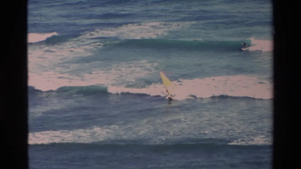 A distant windsurfer in 1981 Hawaii appears near the lower edge of the 8mm frame, surrounded by white surf and blue ocean swell.