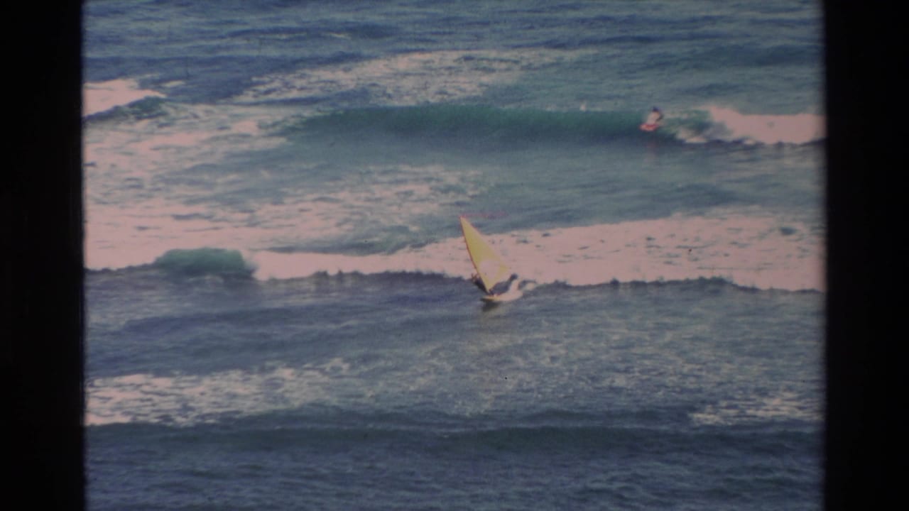 Distant windsurfer with a yellow sail crossing blue ocean waves as a surfer appears near the breaking surf.