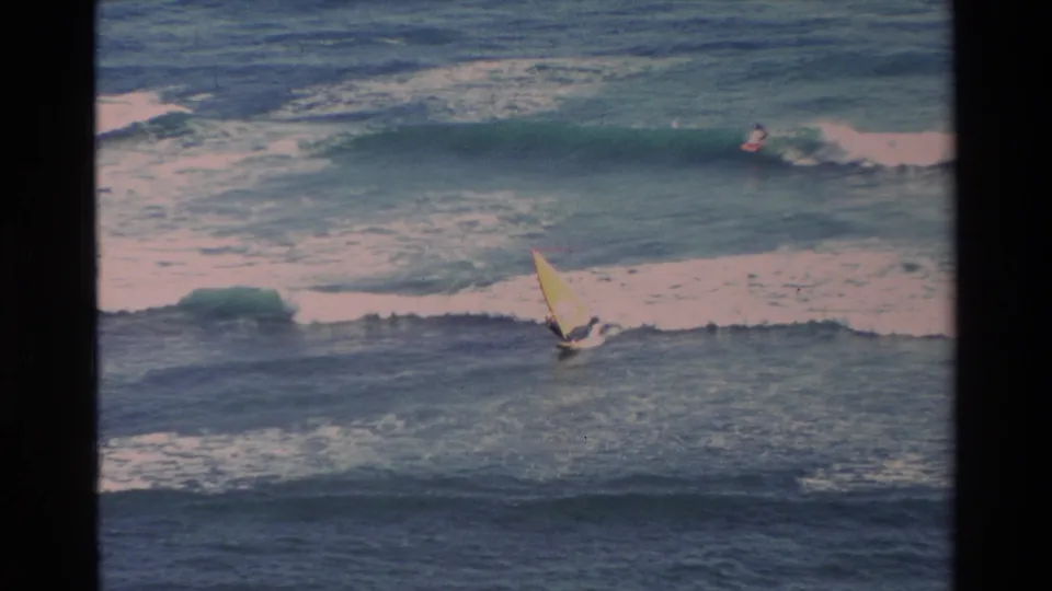 1981 Hawaii 8mm frame showing a yellow-sailed windsurfer on choppy ocean water with a distant surfer near a breaking wave.