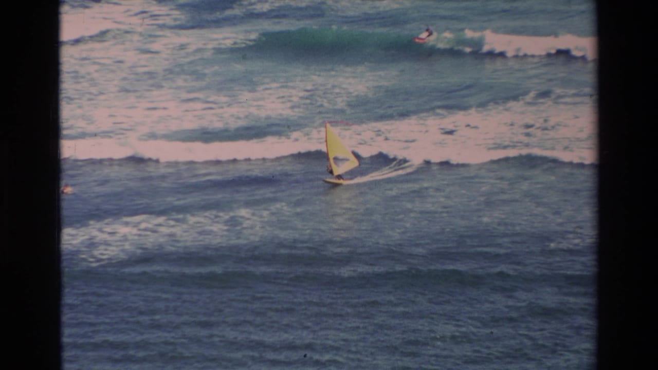 1981 Hawaii home-movie frame of a windsurfer with a yellow sail moving across rough surf as a distant surfer rides behind.