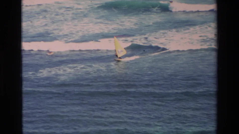 1981 Hawaii 8mm frame showing the yellow windsurf sail closer in the lower surf, with foamy waves filling the background.