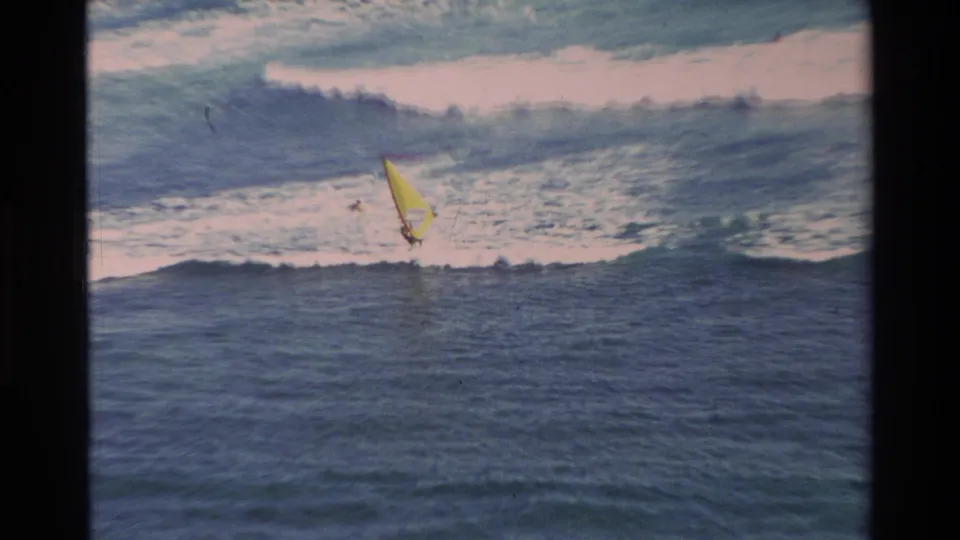 1981 Hawaii home-movie frame of a distant yellow-sailed windsurfer near breaking surf, framed by grain and dark film edges.