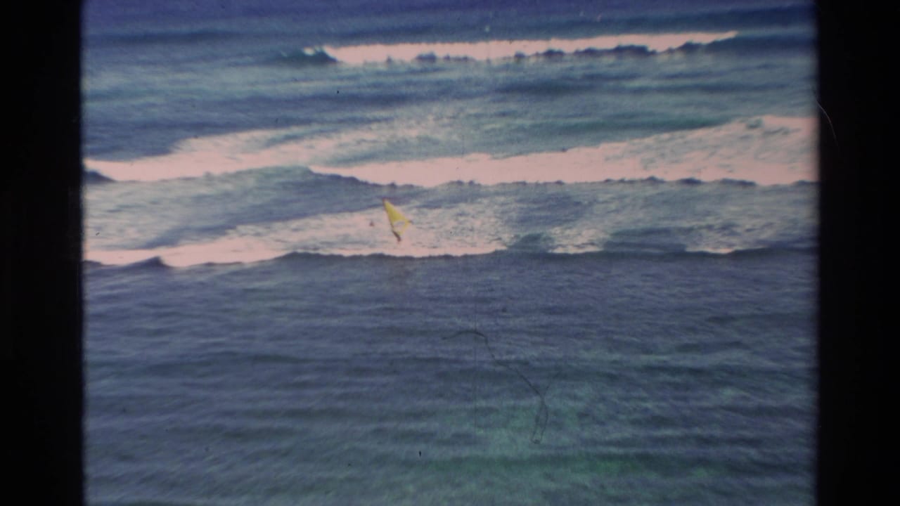 1981 Hawaii 8mm frame showing choppy blue ocean, breaking surf, and a small yellow windsurfing sail in the distance.