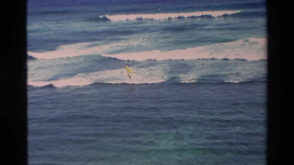 1981 Hawaii 8mm frame showing choppy blue ocean, breaking surf, and a small yellow windsurfing sail in the distance.