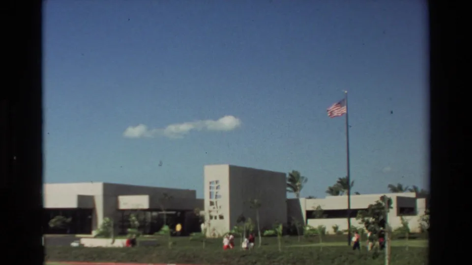 Wide 1981 Hawaii view of the Arizona Memorial Visitor Center grounds with low buildings, lawn, visitors, and an American flag on a tall pole.