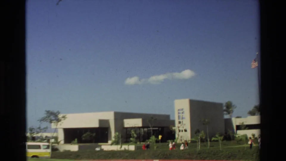 Wide 1981 Hawaii frame showing the visitor center building, small groups of visitors, a parked vehicle, lawn, palms, and clear blue sky.