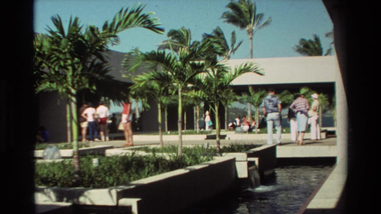 1981 Hawaii 8mm frame showing people gathered by palm trees, a modern resort building, walkway, and water feature in bright sun.