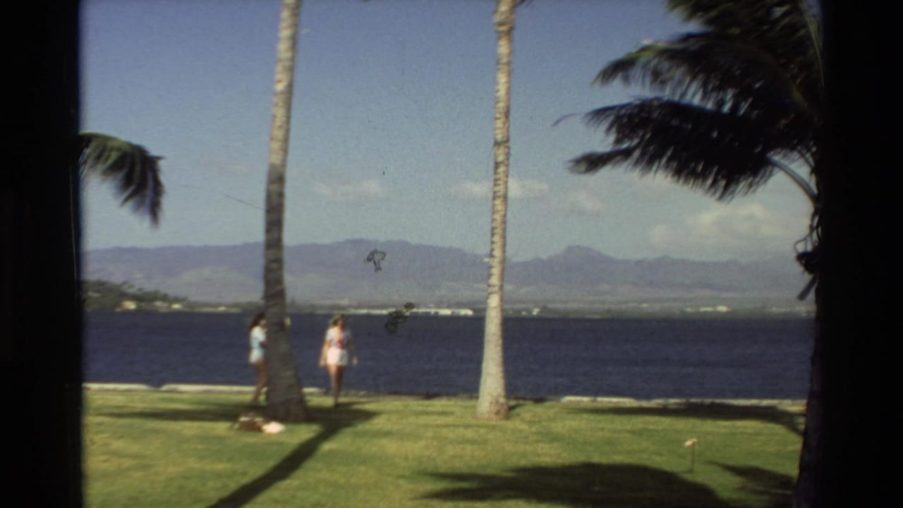 Two women continue walking by the water between tall palm trunks.