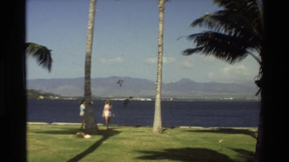 1981 Hawaii frame with two women in light clothing walking along a grassy shoreline framed by palm trees and distant hills.