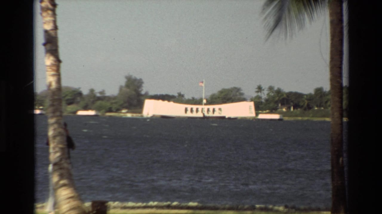 1981 Hawaii home-movie view of the USS Arizona Memorial across Pearl Harbor water, framed by palms and shoreline greenery.