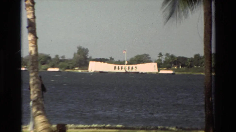 1981 Hawaii home-movie view of the USS Arizona Memorial across Pearl Harbor water, framed by palms and shoreline greenery.