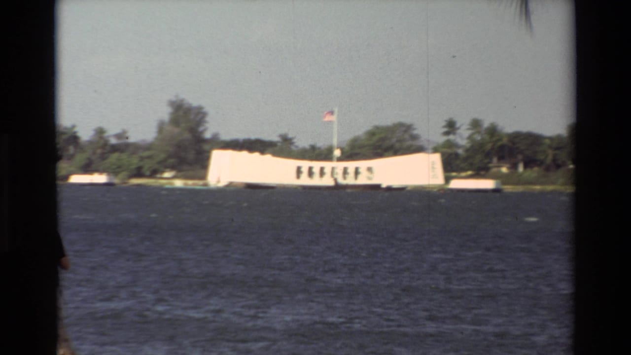 Across-water view of the USS Arizona Memorial in Hawaii, with a U.S. flag and palm-lined shoreline behind it.