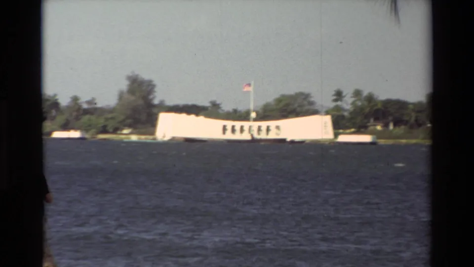 Across-water view of the USS Arizona Memorial in Hawaii, with a U.S. flag and palm-lined shoreline behind it.