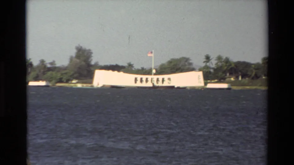 1981 visitor-shot 8mm frame of the USS Arizona Memorial across rippled water with palms and trees behind it.