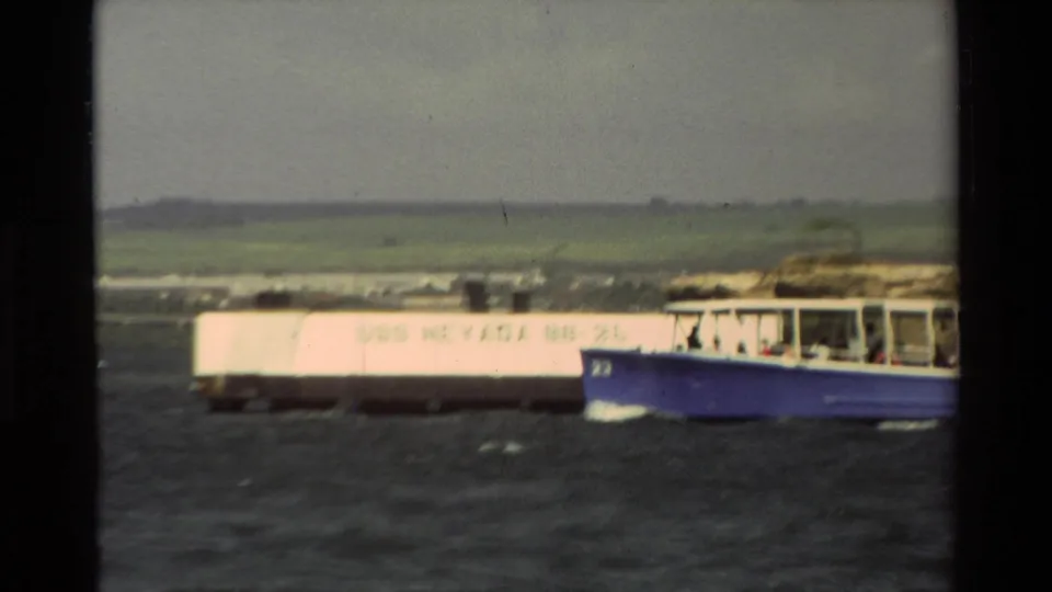 1981 Hawaii home-movie frame with a blue passenger boat passing a pale waterfront structure and distant green hills.