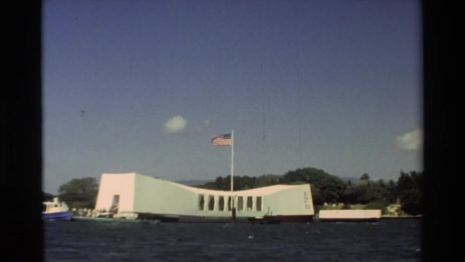 Visitor-shot 1981 Hawaii view of the USS Arizona Memorial, flagpole, shoreline trees, and water in the foreground.