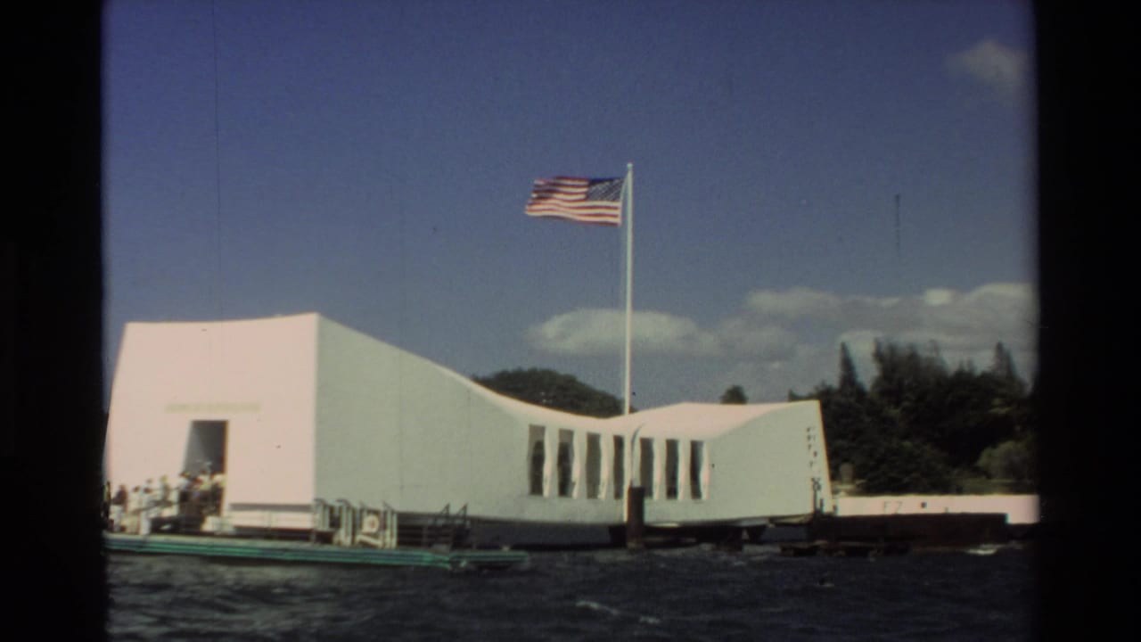 The USS Arizona Memorial appears from a moving boat in Hawaii, with a flagpole, blue sky, clouds, water, and small figures on deck.