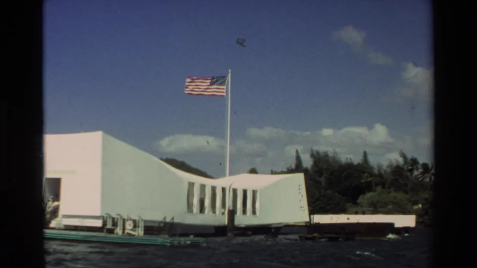 A 1981 8mm frame shows the USS Arizona Memorial from the water, with its white angular facade, flag, blue sky, and green shore behind.