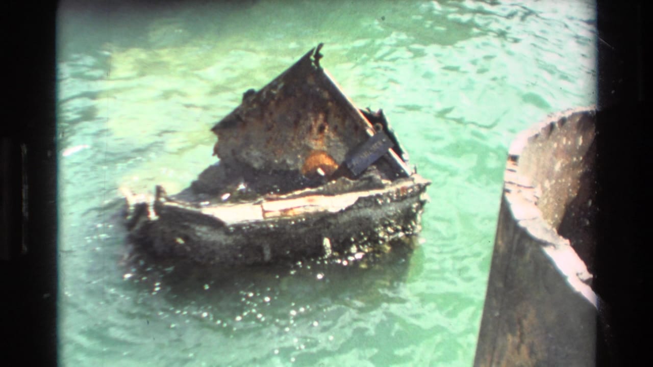 Small weathered wooden boat appears battered in green Hawaiian water, close to a larger dark vessel or dock edge.