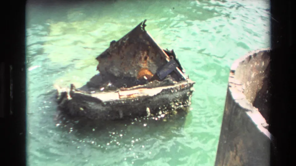 Small weathered wooden boat appears battered in green Hawaiian water, close to a larger dark vessel or dock edge.