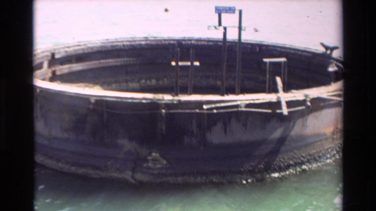 Weathered circular dock or platform floating in clear water with metal railings and dark film edges.
